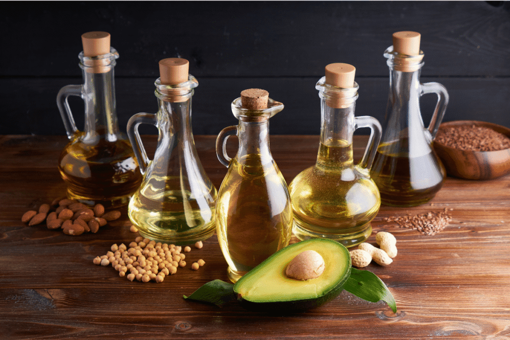 Pflanzenöl: Verschiedene pflanzliche Öle in Glasflaschen – darunter Avocadoöl, Sojaöl, Mandelöl, Leinöl und Erdnussöl – auf einem Holztisch mit passenden Samen und einer Avocado.
