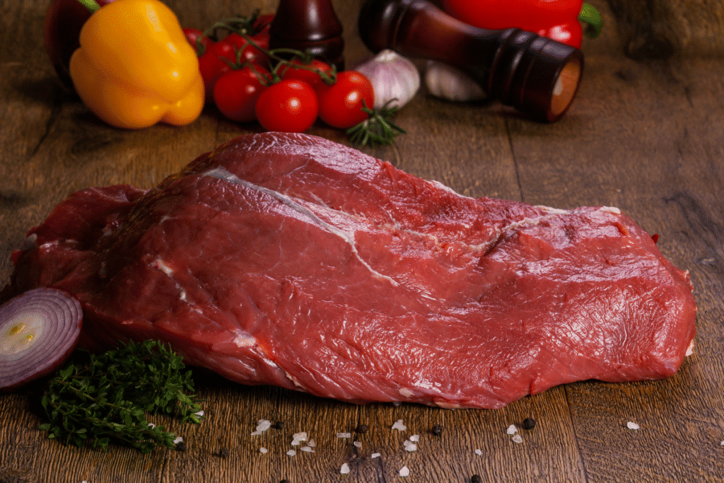 Rotes Fleisch – rohes Rindfleisch auf Holzbrett Rohes Rindfleisch auf Holzbrett, daneben rote Zwiebel und Kräuter; Paprika und Tomaten im Hintergrund