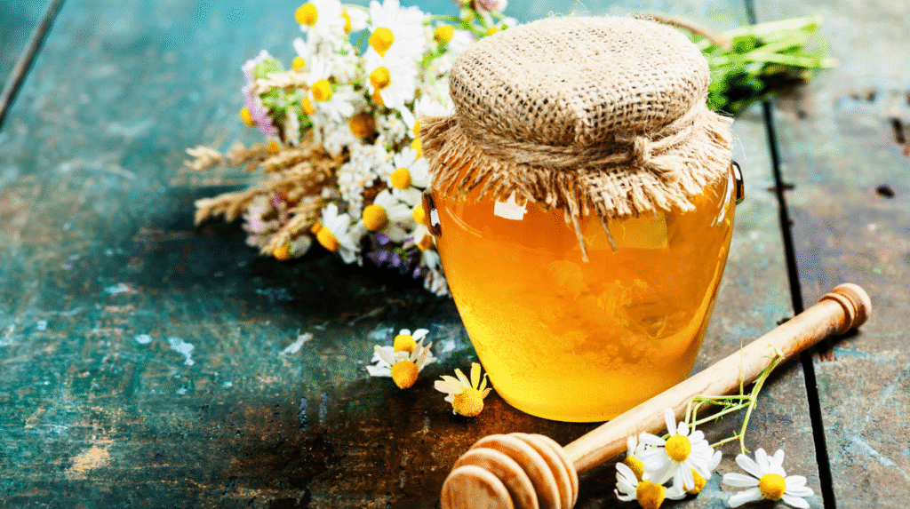 Ist Honig gesund? Glas Honig mit Holzlöffel und Kamillenblüten auf rustikalem Tisch