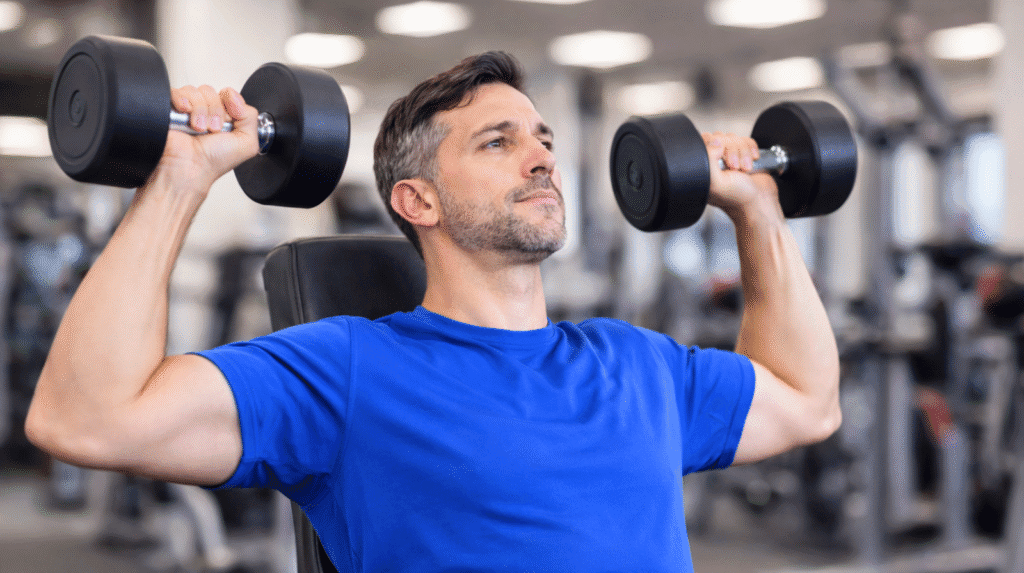 Mann Ende 40 trainiert im Fitnessstudio mit Kurzhanteln beim Schulterdrücken